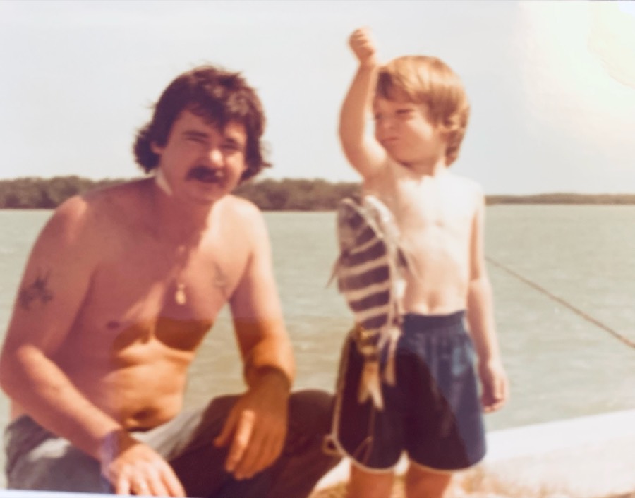 Donald Barde sitting on a dock beside his young son Brian, who is holding up a fish he caught, in the early 1980s