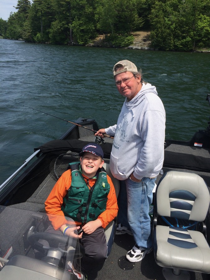 Donald Barde standing in a boat holding a fishing rod behind his grandson, who is seated in an orange jacket and life vest, laughing